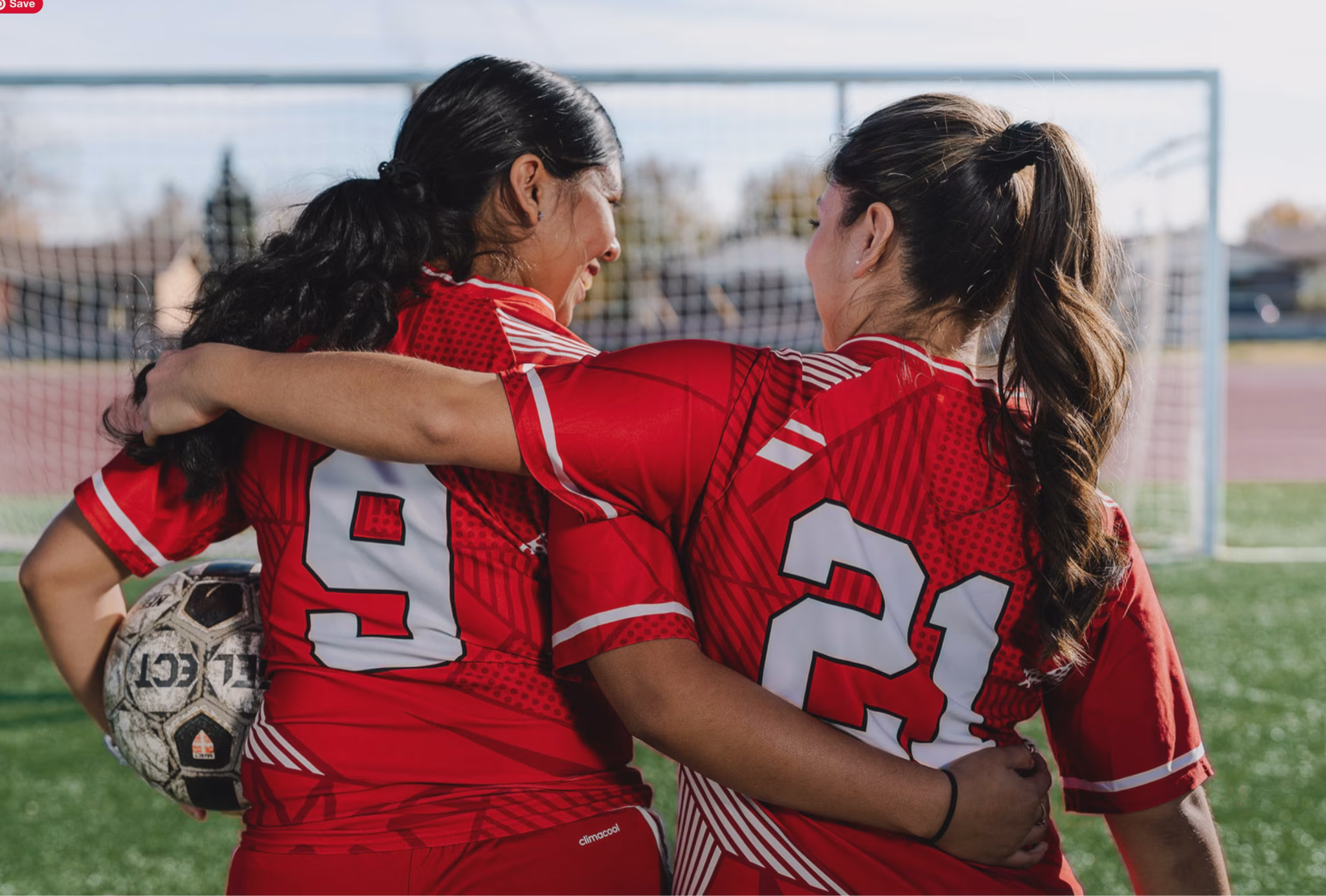 Two female soccer players in red jerseys with their arms around each other and their backs to the camera